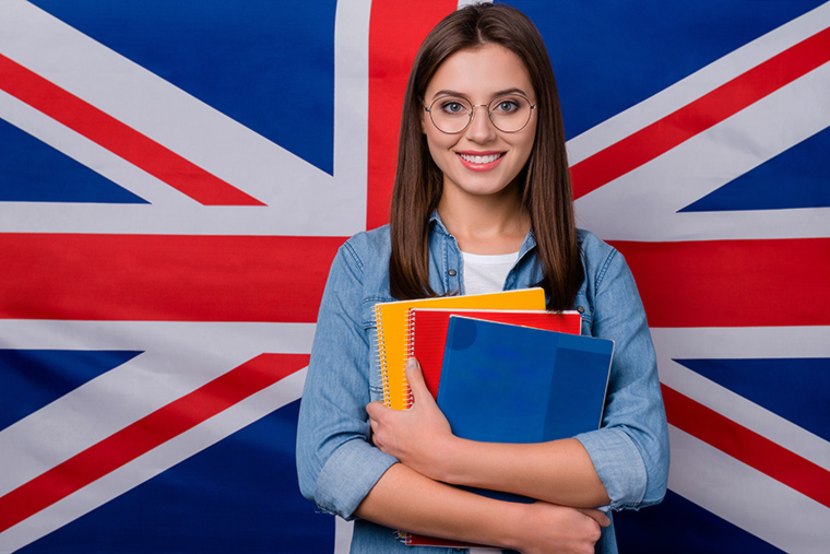 Zbliżony portret ładnej, wesołej dziewczyny w okularach trzymającej w rękach zeszyt. Dziewczyna stoi na tle brytyjskiej flagi. 