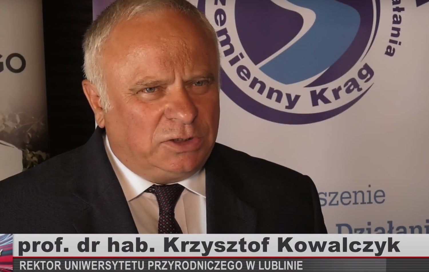 wywiad tv z Rekroerm Krzysztofem Kowalczykiem, w dole podpis, w tle logo partnera uczelni