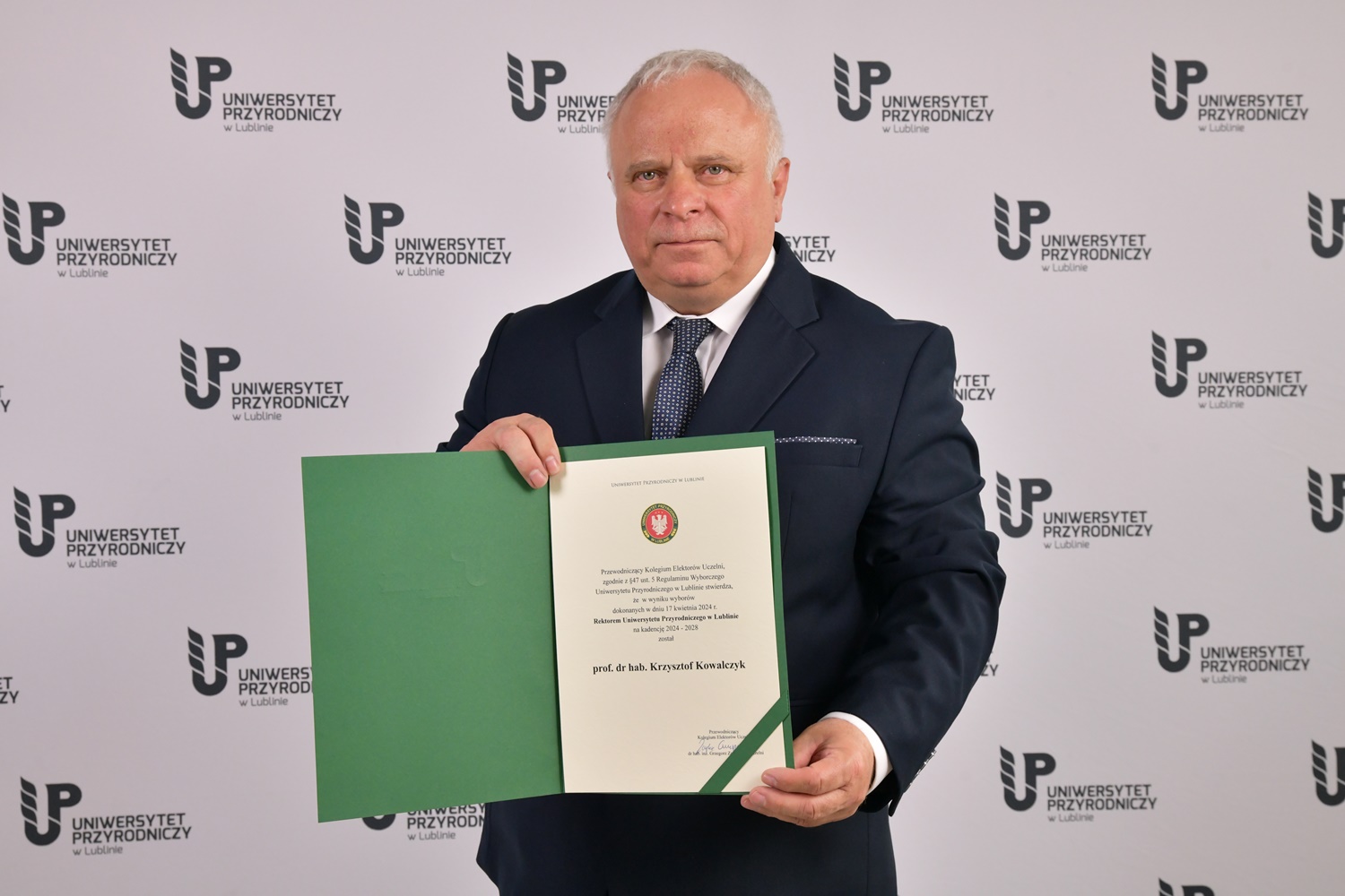 Rektor Krzysztof Kowalczyk z aktem mianowania w tle biała ścianka z logo Uniwersytetu Przyrodniczego w Lublinie