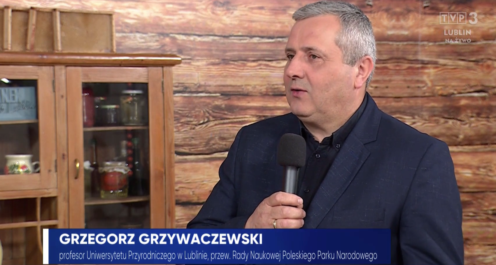 dr hab. Grzegorz Grzywaczewski, prof. uczelni w ciemnym garniturze, z mikrofonem, tle drewniana ściana