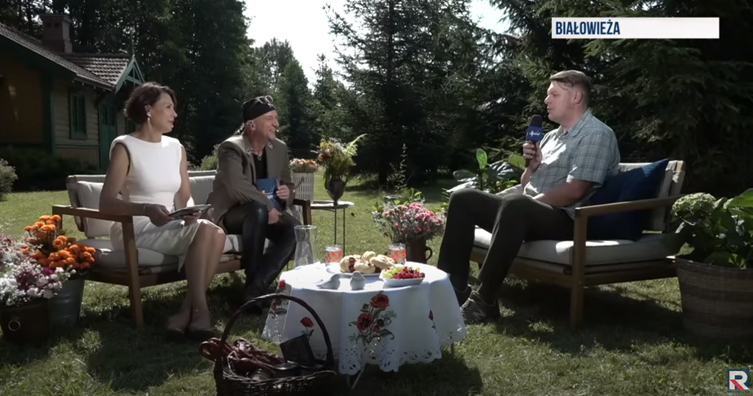 plener, rozmowa 3 osoby: po lewej kobieta i mężczyzna siedzą na kanapie, w środku stół z owo i bułkami, po prawej: prof. Michał Krzysiak 