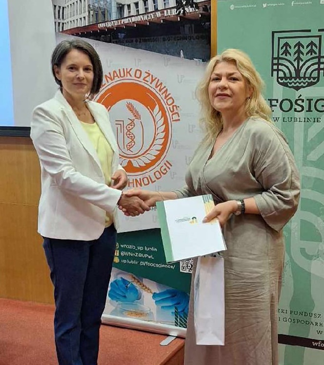 Dr inż. Monika Michalak-Majewska (ciemne włosy, biały żakiet, granatowe dżinsy), wręcza powinszowanie uczestniczce warsztatów (jasne włosy. kremowa sukienka)