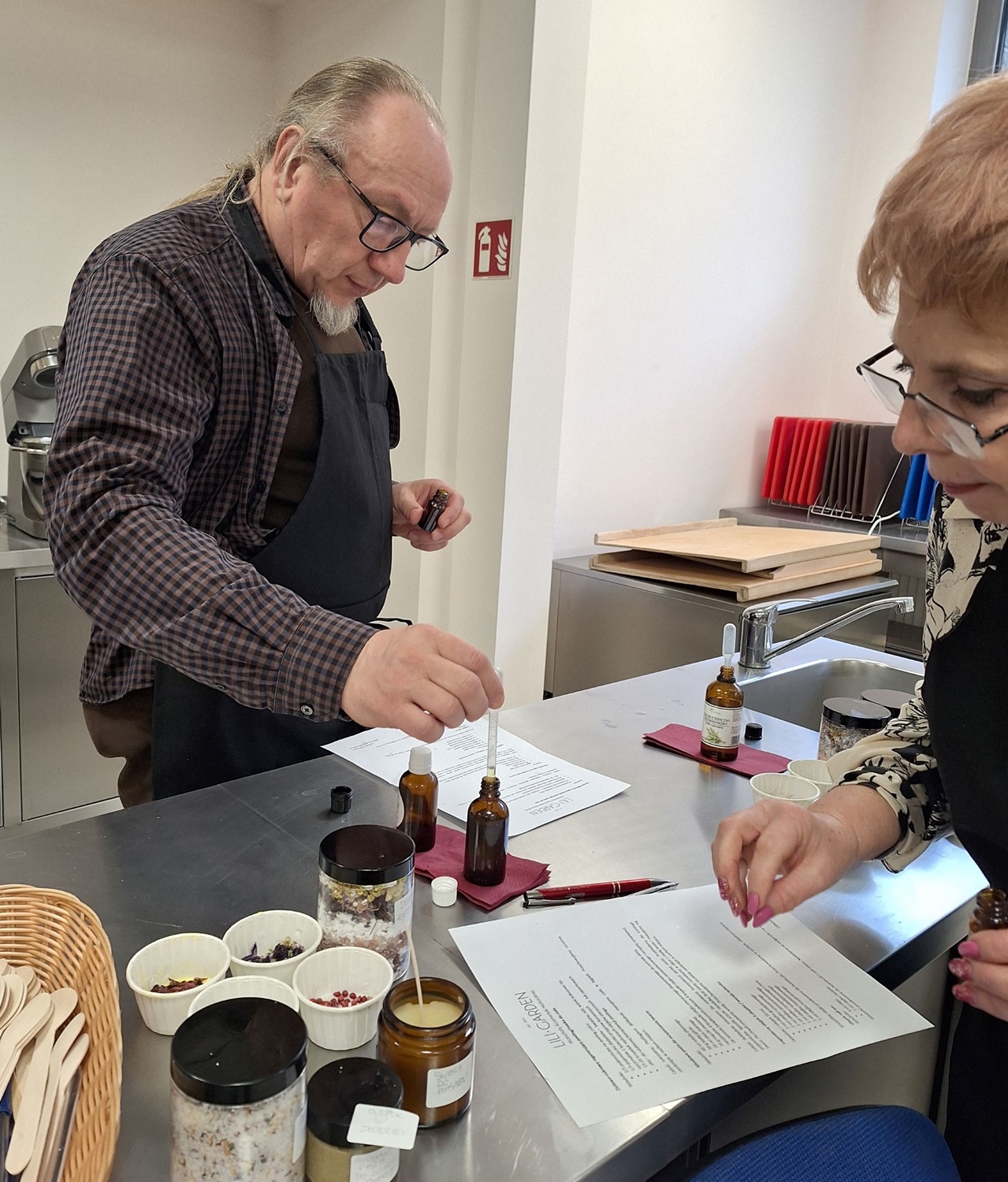 mężczyzna w ciemnej koszuli tworzy kosmetyki po prawej kobieta w czerni robi notatki