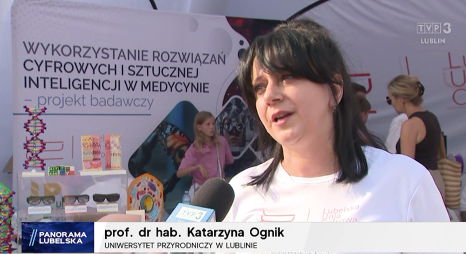 prof. Katarzyna Ognik, biały t-shirt, czarne włosy do ramion, w tle banery projektu UP w Lublinie