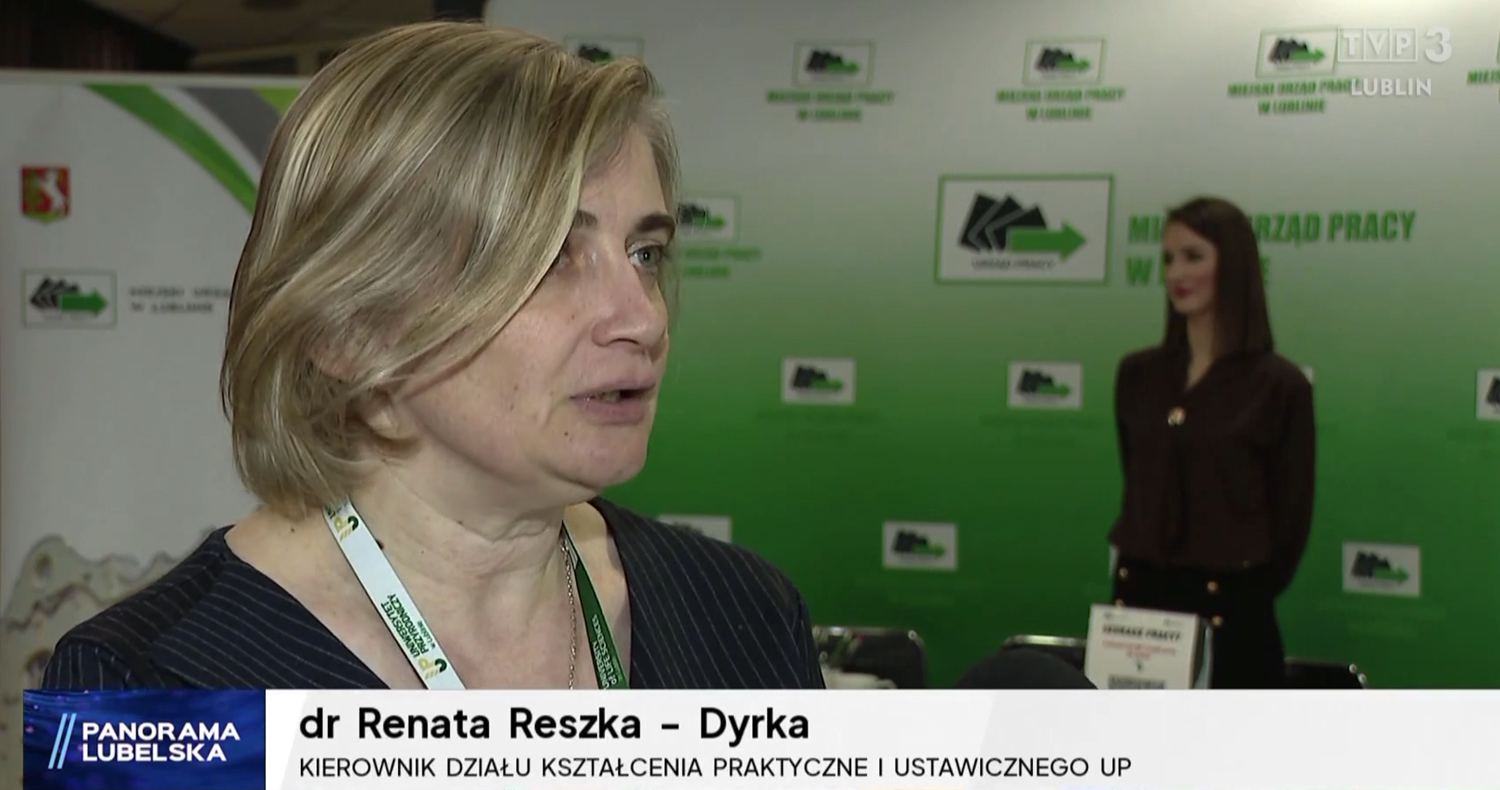 dr Renata Reszka-Dyrka, krótkie blond włosy, ciemnoszary żakiet w&nbsp;szare paski, w&nbsp;tle zielona ścianka i&nbsp;stojąca kobieta ubrana na ciemno