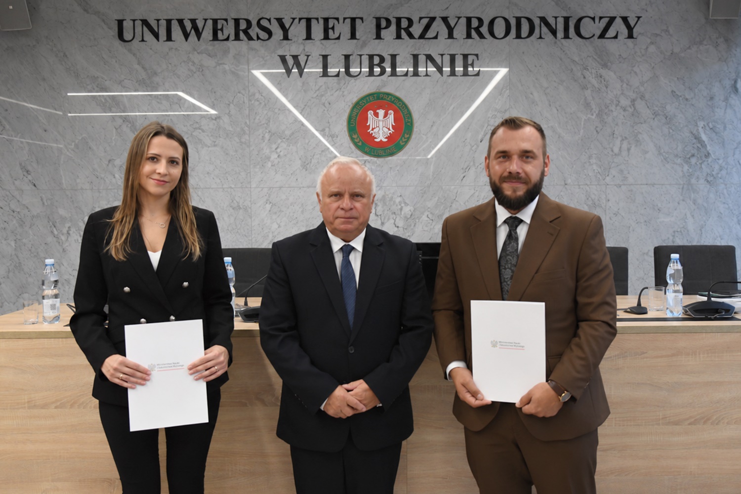 Rektor prof. dr hab. Krzysztof Kowalczyk w&nbsp;czarnym garniturze, obok dr inż. Małgorzata Sierocka w&nbsp;czarnym spodium i&nbsp;dr inż. Kamil Drabik w&nbsp;brązowym garniturze, na tle granitowej ściany z&nbsp;napisem Uniwersytet Przyrodniczy w&nbsp;Lublinie i&nbsp;czerwono-zielonym godłem z&nbsp;białym orłem