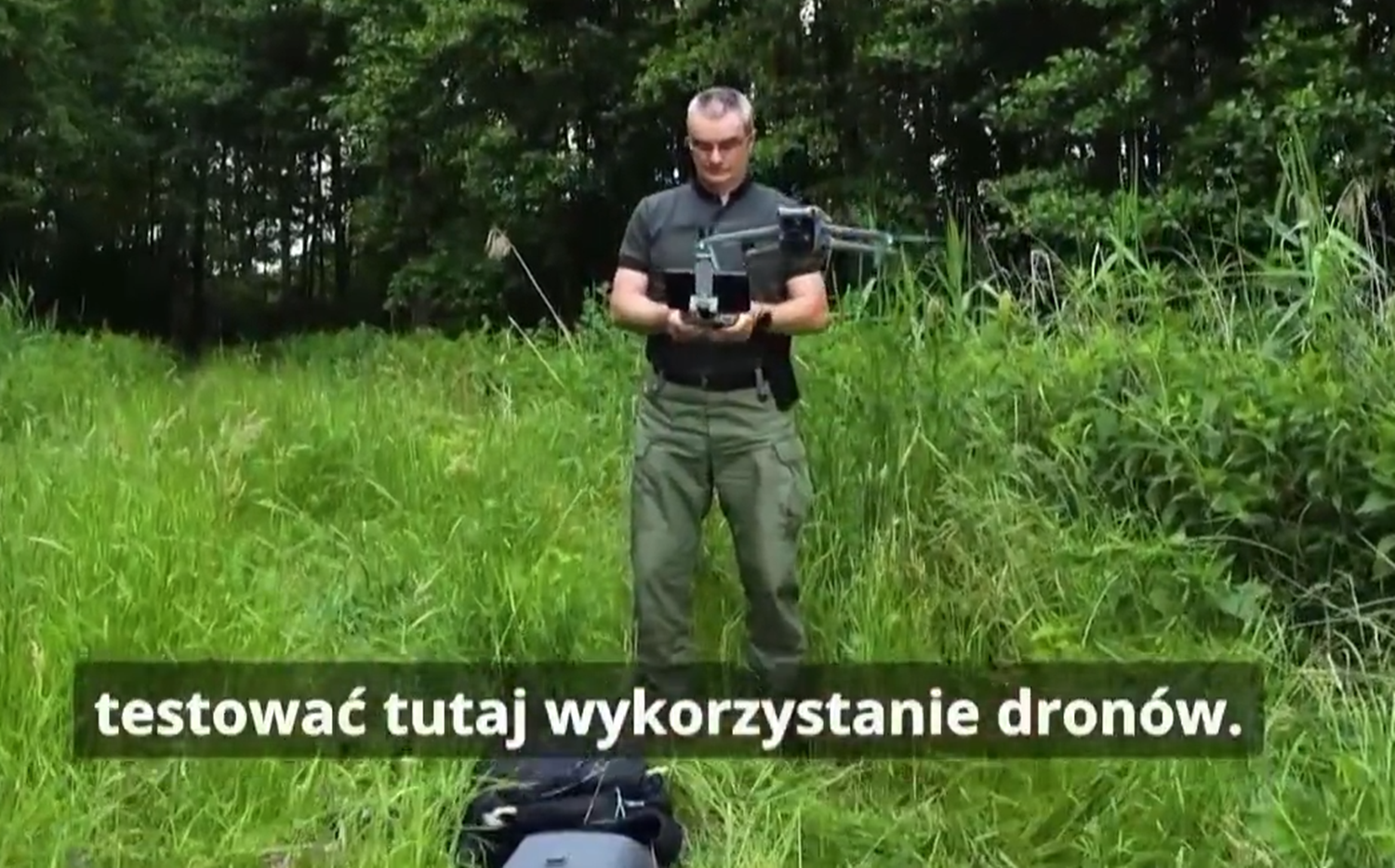 dr Mariusz Wójcik w&nbsp;czarnej koszuli i&nbsp;spodniach khaki stoi na tle zieleni i&nbsp;steruje dronem